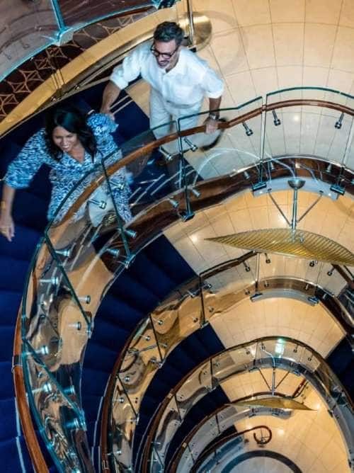 Couple ascends an atrium staircase aboard an all-inclusive, luxury Seabourn cruise.