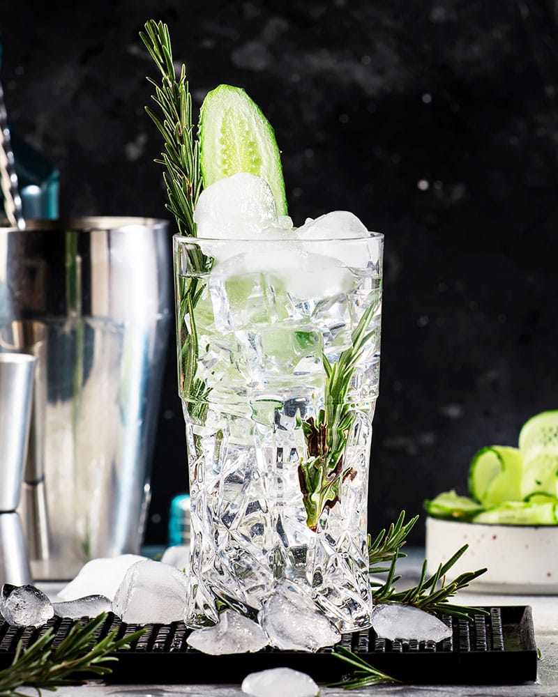 Gin tonic with cucumber, alcoholic long drink cocktail with dry gin, rosemary, tonic, fresh cucumber and ice cubes. Black background, bar tools, copy space