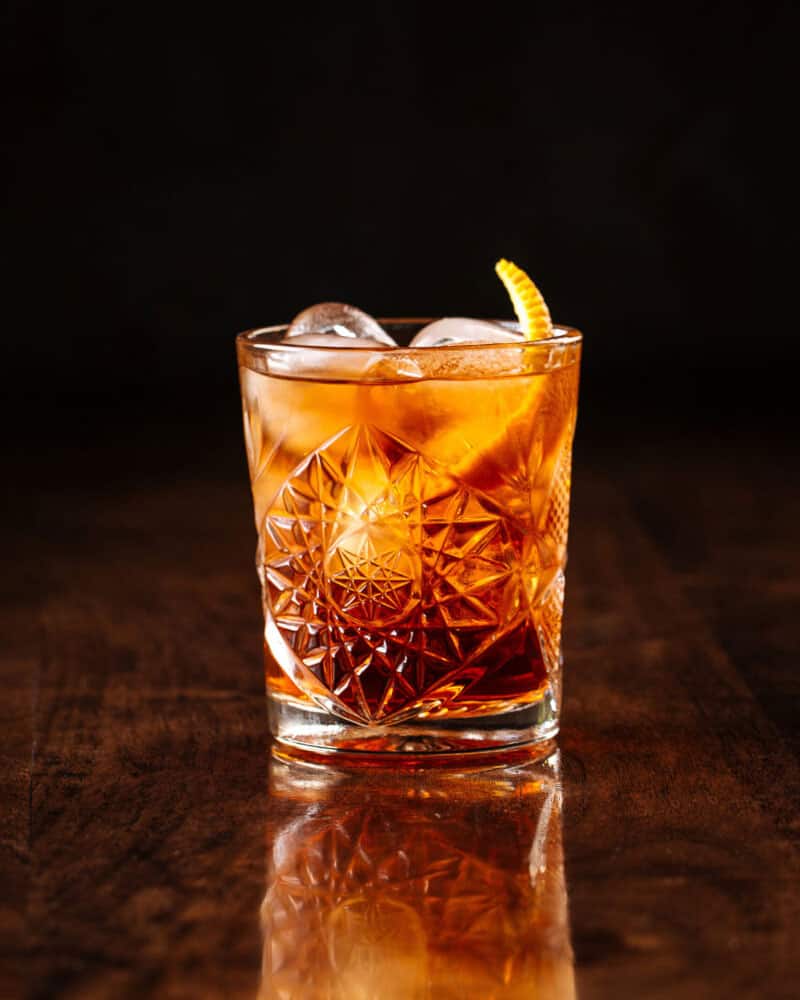 delicious old fashion cocktail in the etched glass with ice and orange slices, dark wooden background, vertical, side view