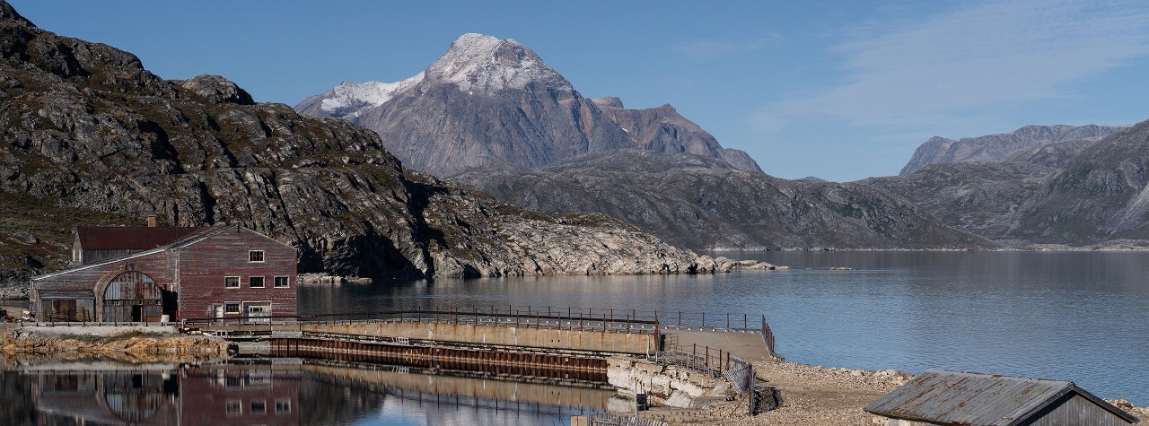 Rocky Greenland fjord with calm water, a weathered red wooden building and pier on shore, backed by rugged mountains under clear skies.