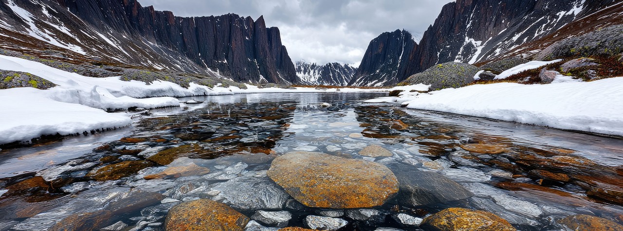 Clear stream flows between snow-lined banks into a rugged fjord, framed by steep dark cliffs and spring snow under cloudy skies. Rocky stream, spring melt, Torngat Mountains, Canada