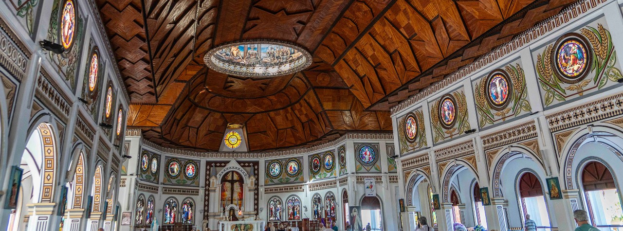 Inside an ornate island church, carved wood ceiling arches over stained‑glass windows and decorative columns visited by Seabourn guests.