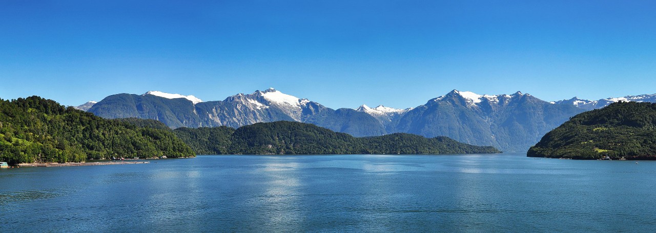 A calm blue fjord framed by forested hills and snow‑capped mountains under a clear sky during a Seabourn shore excursion.