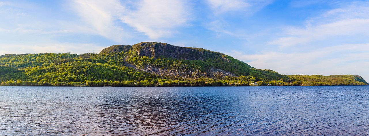 Calm blue channel bordered by forested green hills and a rocky ridge under a clear sky, showcasing a scenic waterway.
