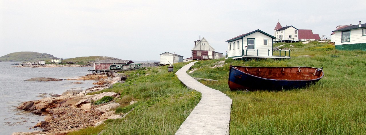 Wooden boardwalk winds through a quiet Labrador coastal village with colorful houses, grassy shoreline, and small boats on a Seabourn shore excursion. Hopedale