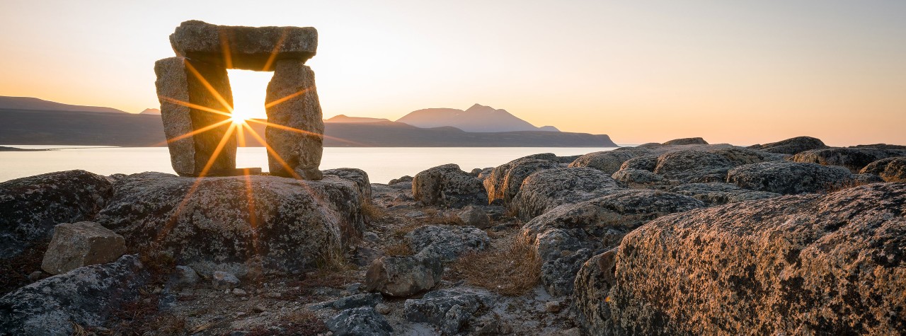 Sunlight shines through a small stone arch on a rocky Arctic shoreline, with calm water and distant mountains at golden hour. Sunset through Inuksuk in rough arctic landscape. Sunstar on the horizon in the fjord of Qikiqtarjuaq, Broughton Island, Nunavut.