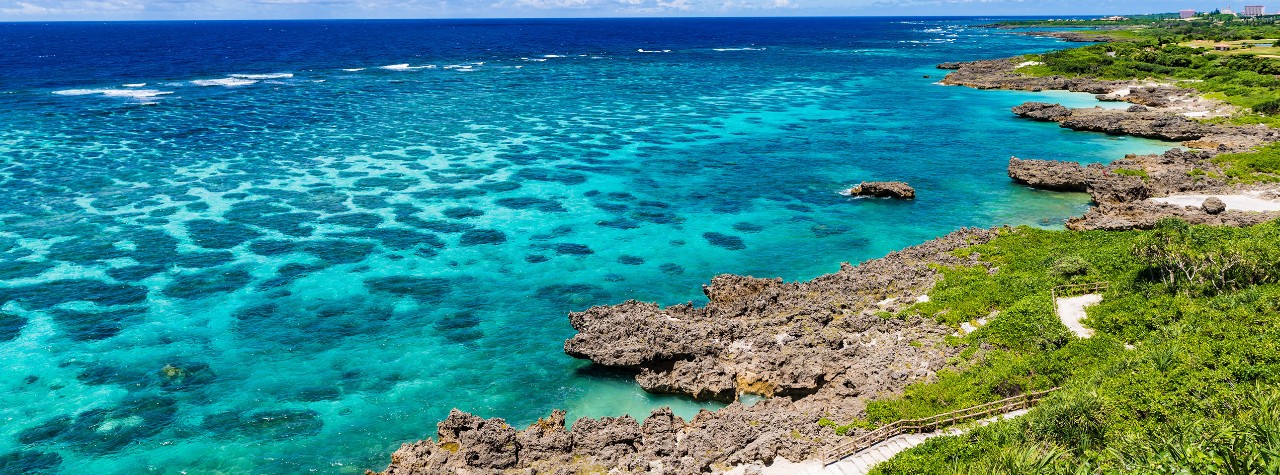Turquoise reef-filled waters along a rocky, green coastline in Miyakojima, Japan, with waves breaking offshore during a scenic Seabourn shore excursion.