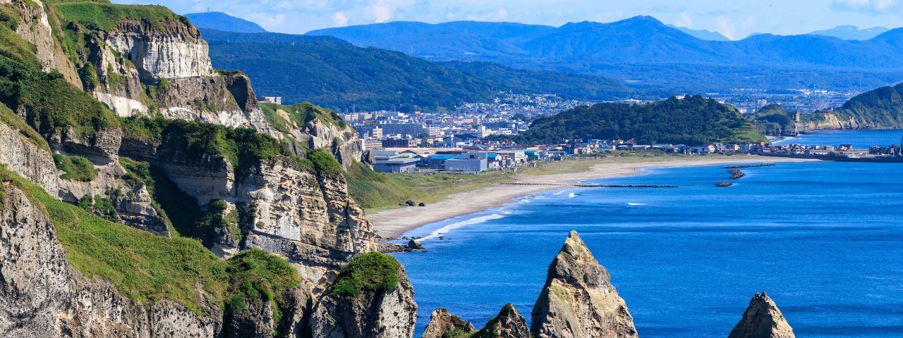 Jagged rocks hugging coast with mountains and city in background in Muroran, Japan