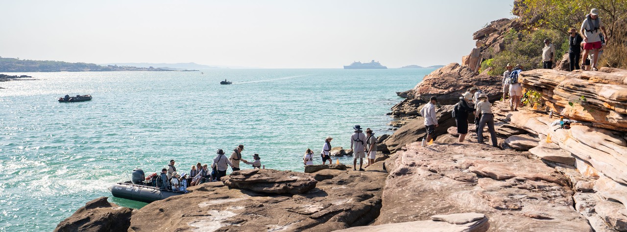 Seabourn guests explore a rocky Swift Bay shoreline during a shore excursion, with Zodiac boats nearby and an ultra-luxury Seabourn ship offshore.