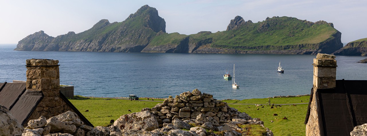Stone ruins overlook a green coastal meadow and sheltered bay with small boats, backed by rugged island cliffs in St Kilda.