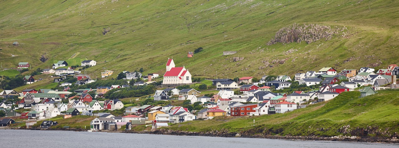 Colorful hillside village in the Faroe Islands with a red-roofed church, clustered homes, and green slopes above a calm shoreline. Traditional faroese village in Suduroy island. Fjord landscape. Tvoroyri